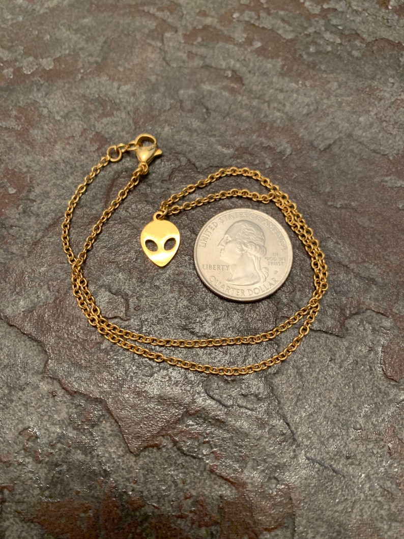 May include: A gold chain necklace with a small alien head pendant. The necklace is laying on a textured surface next to a US quarter.