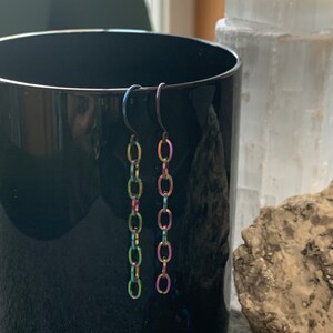 May include: A pair of iridescent chain earrings with a black hook closure. The earrings are made of metal and have a rainbow finish.
