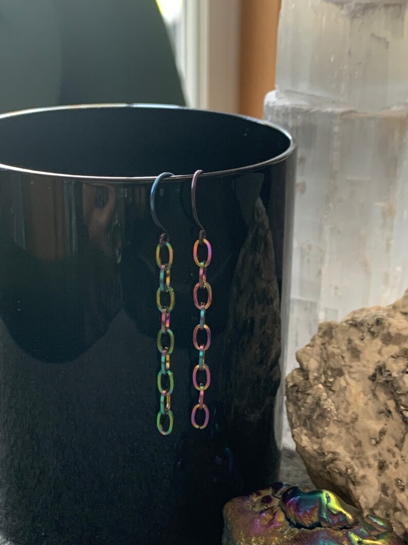 May include: A pair of iridescent chain earrings with a black hook closure. The earrings are made of metal and have a rainbow finish.
