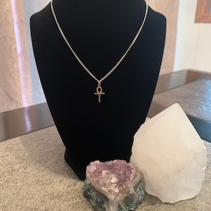 May include: A silver chain necklace with an ankh pendant. The necklace is displayed on a black mannequin bust. There are two crystals, one white and one purple, on the table below the bust.