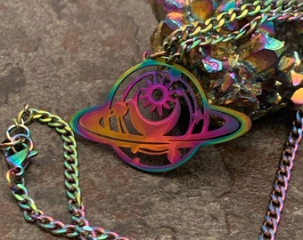 May include: A rainbow iridescent chain necklace with a pendant featuring a stylized sun, moon, and planets. The pendant is made of metal and has a cutout design.