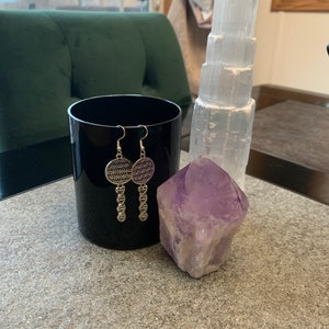 May include: A pair of silver earrings with a flower of life design dangle from a chain. The earrings are displayed on a black surface next to a clear selenite tower and a purple amethyst crystal.