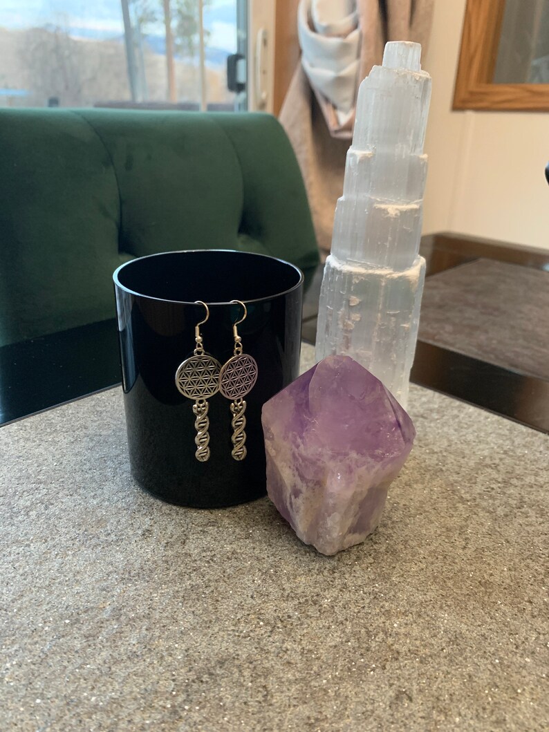 May include: A pair of silver earrings with a flower of life design dangle from a chain. The earrings are displayed on a black surface next to a clear selenite tower and a purple amethyst crystal.