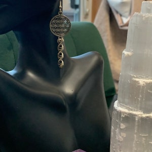 May include: Silver earrings featuring a flower of life design and a dangling chain. The earrings are displayed on a black mannequin bust.