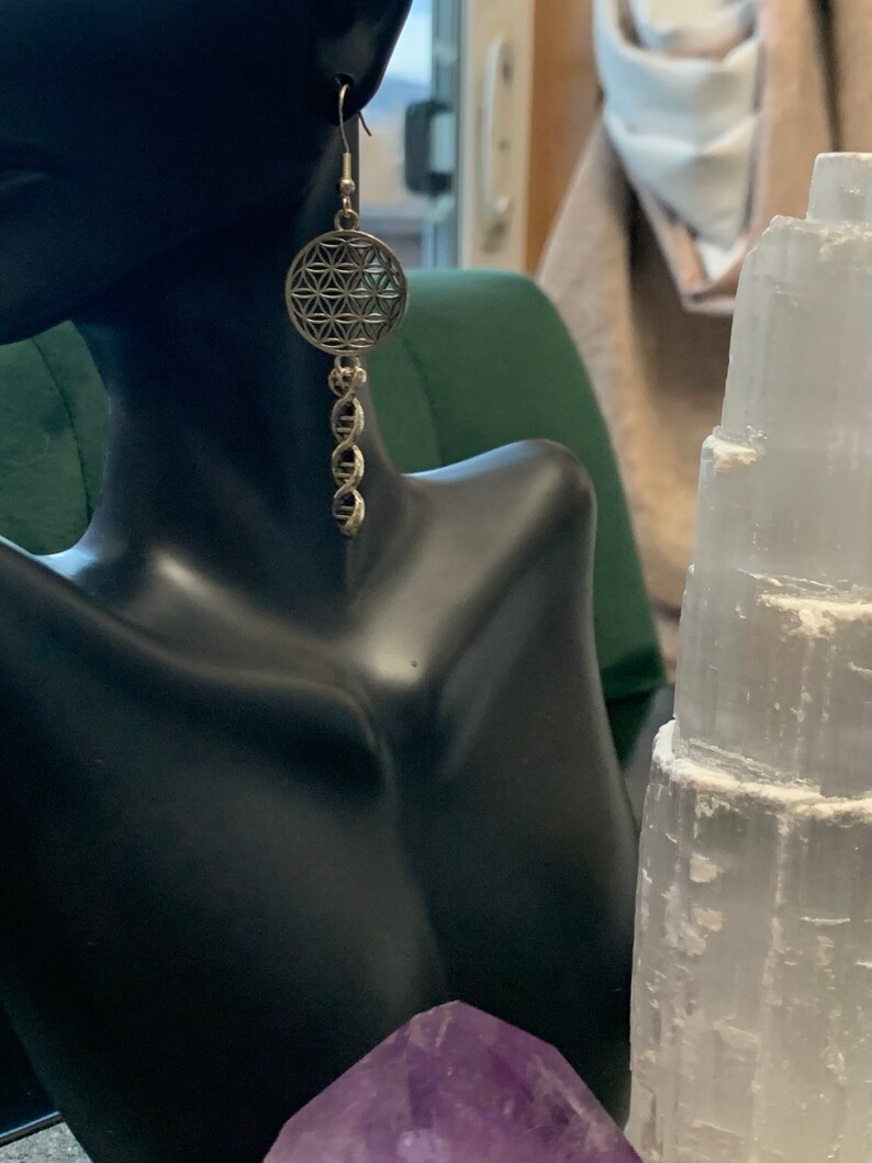 May include: Silver earrings featuring a flower of life design and a dangling chain. The earrings are displayed on a black mannequin bust.