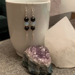 May include: A pair of silver earrings with black and white beads. The earrings are hanging from a white mug and a white crystal.