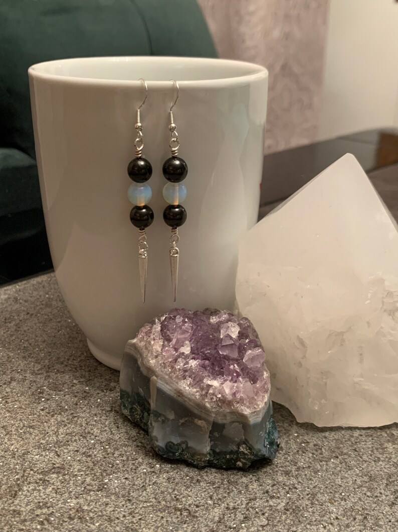 May include: A pair of silver earrings with black and white beads. The earrings are hanging from a white mug and a white crystal.