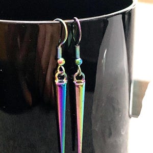 May include: A pair of iridescent silver earrings with a pointed, teardrop-shaped design. The earrings are hanging from a black surface.
