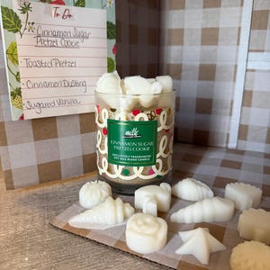 May include: A glass candle jar with white wax melts, labelled "Cinnamon Sugar Pretzel Cookie." Various white wax melt shapes are on a cardboard surface. A handwritten to-do list is visible, with the words "Cinnamon Sugar Pretzel Cookie."