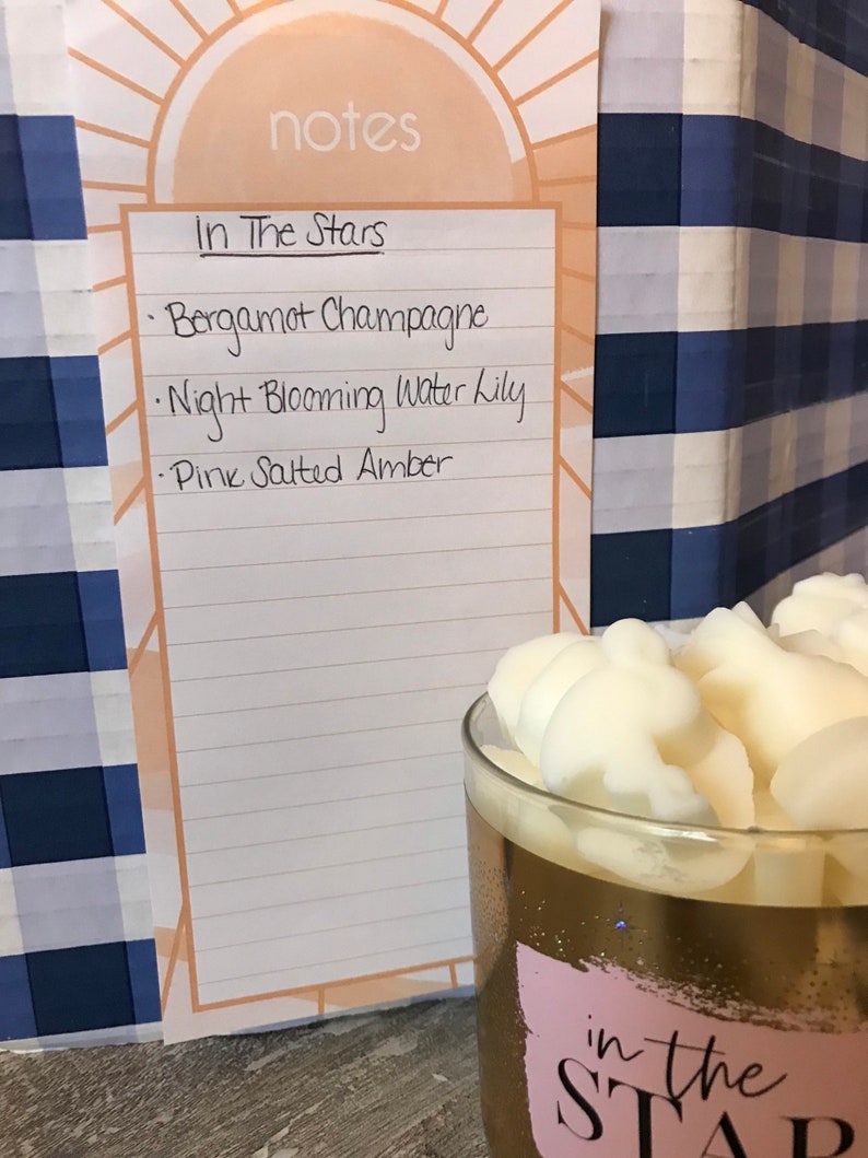 May include: A white notebook with the words "notes" and "In The Stars" written on it. The notebook lists the following scents: Bergamot Champagne, Night Blooming Water Lily, and Pink Salted Amber.