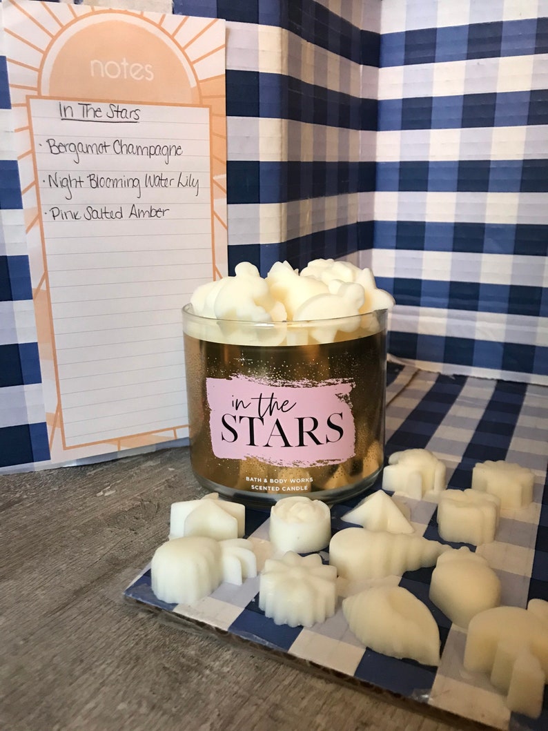 May include: A gold candle jar with the text "in the STARS" and the brand name "BATH & BODY WORKS" on the label. The candle is surrounded by small white wax melts in various shapes. The candle and melts are on a blue and white checkered surface.