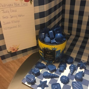 May include: A blue candle jar with the label "Midnight Blue Citrus" and a list of ingredients: Juicy Citrus, Fresh Spring Water, Sweet Mango, and Essential Oils. The candle jar is surrounded by blue wax melts shaped like seashells, pineapples, stars, and other ocean-themed shapes.