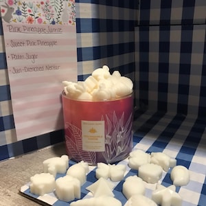 Puede incluir: Un tarro de vela rosa con una etiqueta blanca que dice "Pink Pineapple Sunrise" y una lista de ingredientes: "Sweet Pine Pineapple", "Palm Sugar" y "Sun-Drenched Nectar". El tarro está rodeado de fundidos de cera blanca en varias formas, incluyendo mariposas, estrellas y hojas.