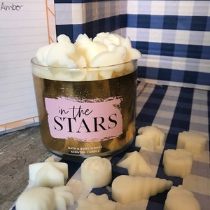 May include: A gold candle jar with the text "in the STARS" and the words "Bath & Body Works Scented Candle". The jar is filled with white wax melts shaped like stars, moons, and other celestial objects. The candle is sitting on a blue and white checkered surface.