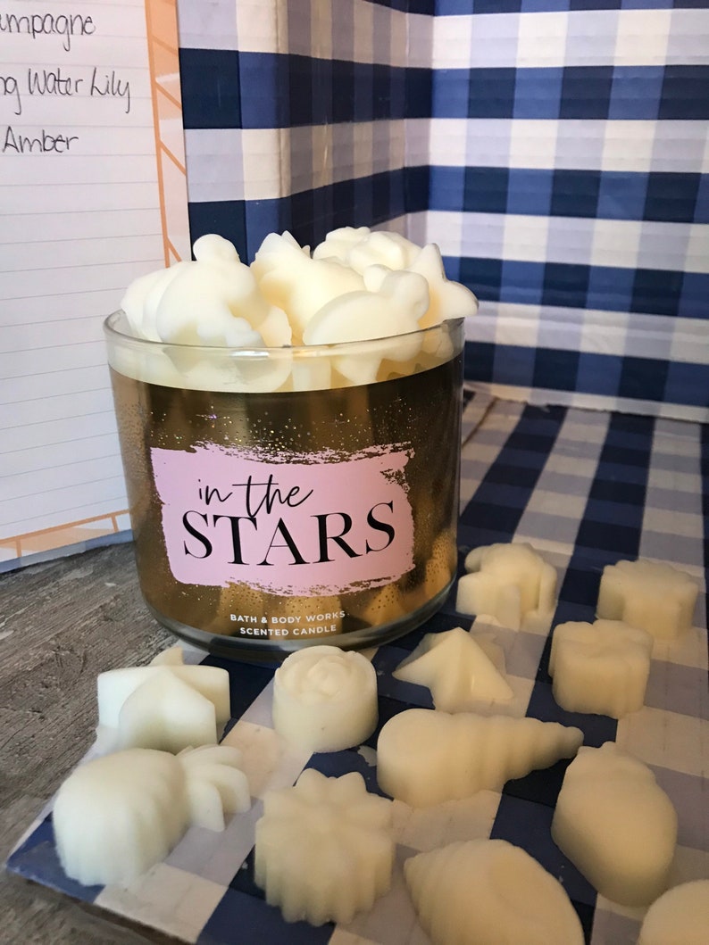 May include: A gold candle jar with the text "in the STARS" and the words "Bath & Body Works Scented Candle". The jar is filled with white wax melts shaped like stars, moons, and other celestial objects. The candle is sitting on a blue and white checkered surface.