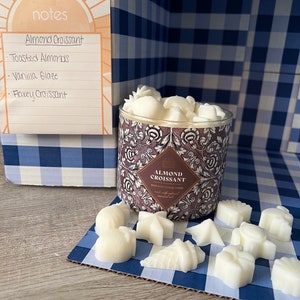 May include: A white candle jar with a brown floral design and the text "Almond Croissant" on the label. The candle is surrounded by small white wax melts in various shapes, including a star, a crescent moon, and a leaf. The wax melts are on a blue and white checked surface.