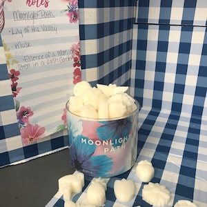 May include: A glass jar filled with white wax melts, labeled "Moonlight Path". The jar is surrounded by a blue and white checkered surface. The wax melts are shaped like flowers, stars, and other designs. The jar is sitting on a blue and white checkered surface. The image also includes a note card with the text "notes" and a list of scents: "Moonlight Path", "Lily of the Valley", "Musk", "Essence of a Moonlight Stroll in a Lush Garden".