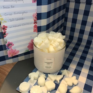 May include: A white jar candle with the text "Laundry Day" on the label. The candle is surrounded by white wax melts in various shapes, including a pineapple, a butterfly, and a flower. The wax melts are on a blue and white checkered surface.