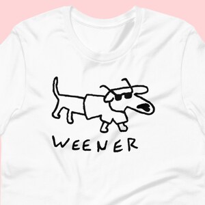 May include: White t-shirt featuring a black line drawing of a dachshund wearing sunglasses and a hat. The text "WEENER" is printed below the dog.