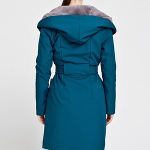 May include: A teal long-sleeved coat featuring a hood lined with grey faux fur. The coat includes a matching fabric belt at the waist, long sleeves, and a smooth, solid-colored exterior finish that extends to the mid-thigh length.