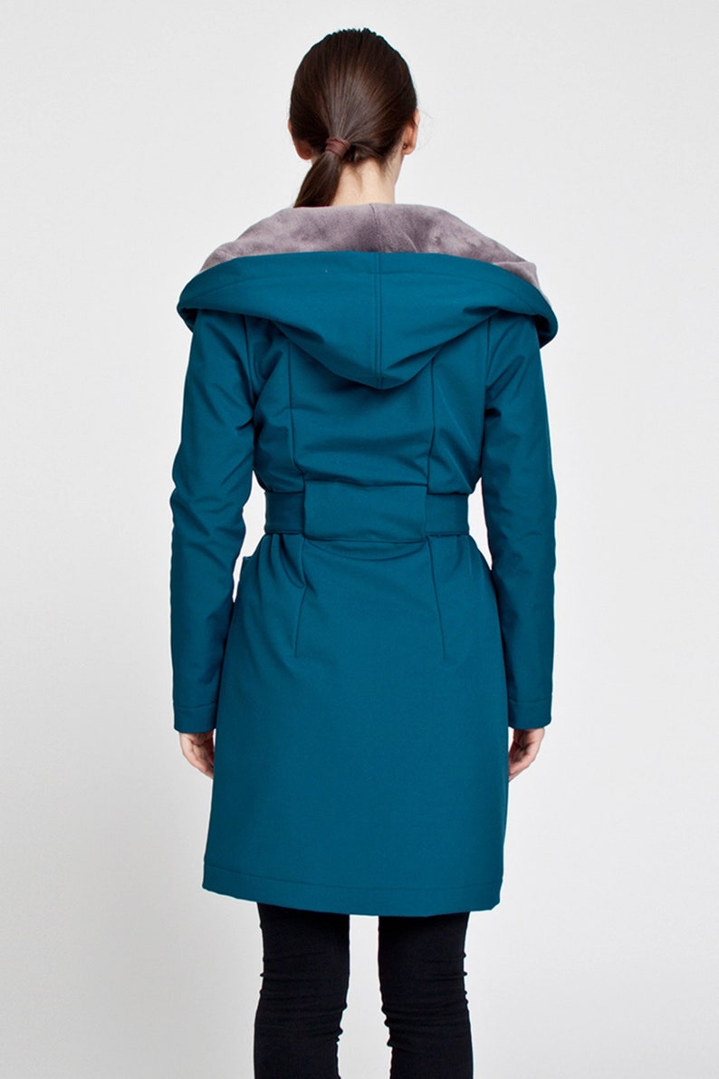 May include: A teal long-sleeved coat featuring a hood lined with grey faux fur. The coat includes a matching fabric belt at the waist, long sleeves, and a smooth, solid-colored exterior finish that extends to the mid-thigh length.