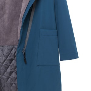 May include: A teal blue coat featuring a hood, a front zipper closure with a black pull tab, and a large patch pocket on the side. The interior is lined with a soft, dark gray fleece upper section and a quilted, diamond-patterned lower section.