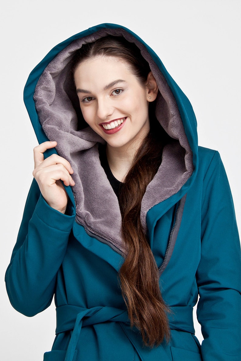May include: A teal hooded coat featuring a soft, grey fleece lining inside the hood. The coat includes a matching teal fabric belt tied at the waist and a full-length front zipper closure, providing a structured look with long sleeves.