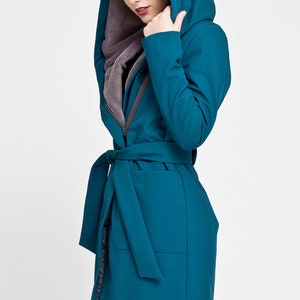 May include: A teal hooded coat featuring a long silhouette, a matching fabric tie belt at the waist, and a contrasting grey interior lining. The garment includes a deep hood and long sleeves, providing a structured look with a smooth exterior finish.