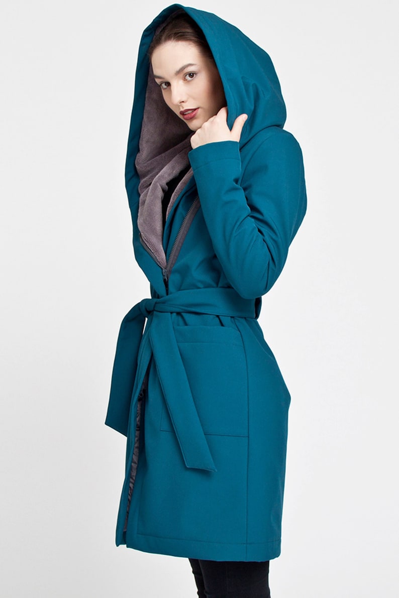 May include: A teal hooded coat featuring a long silhouette, a matching fabric tie belt at the waist, and a contrasting grey interior lining. The garment includes a deep hood and long sleeves, providing a structured look with a smooth exterior finish.