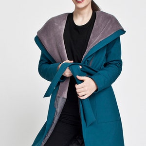 May include: A teal long-sleeved coat featuring a large, wide collar lined with soft grey faux fur. The coat includes a matching fabric tie belt at the waist, two front patch pockets, and a quilted grey interior lining visible at the front opening.