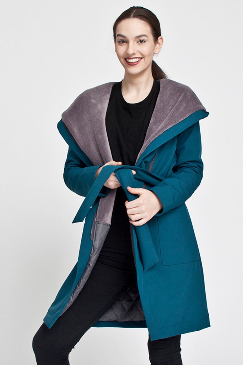 May include: A teal long-sleeved coat featuring a large, wide collar lined with soft grey faux fur. The coat includes a matching fabric tie belt at the waist, two front patch pockets, and a quilted grey interior lining visible at the front opening.