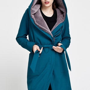 May include: A teal hooded coat featuring a soft grey faux fur lining, a wrap-style front with a matching fabric tie belt, long sleeves, and two front pockets. The coat has an oversized hood and a zipper closure partially visible under the wrap.