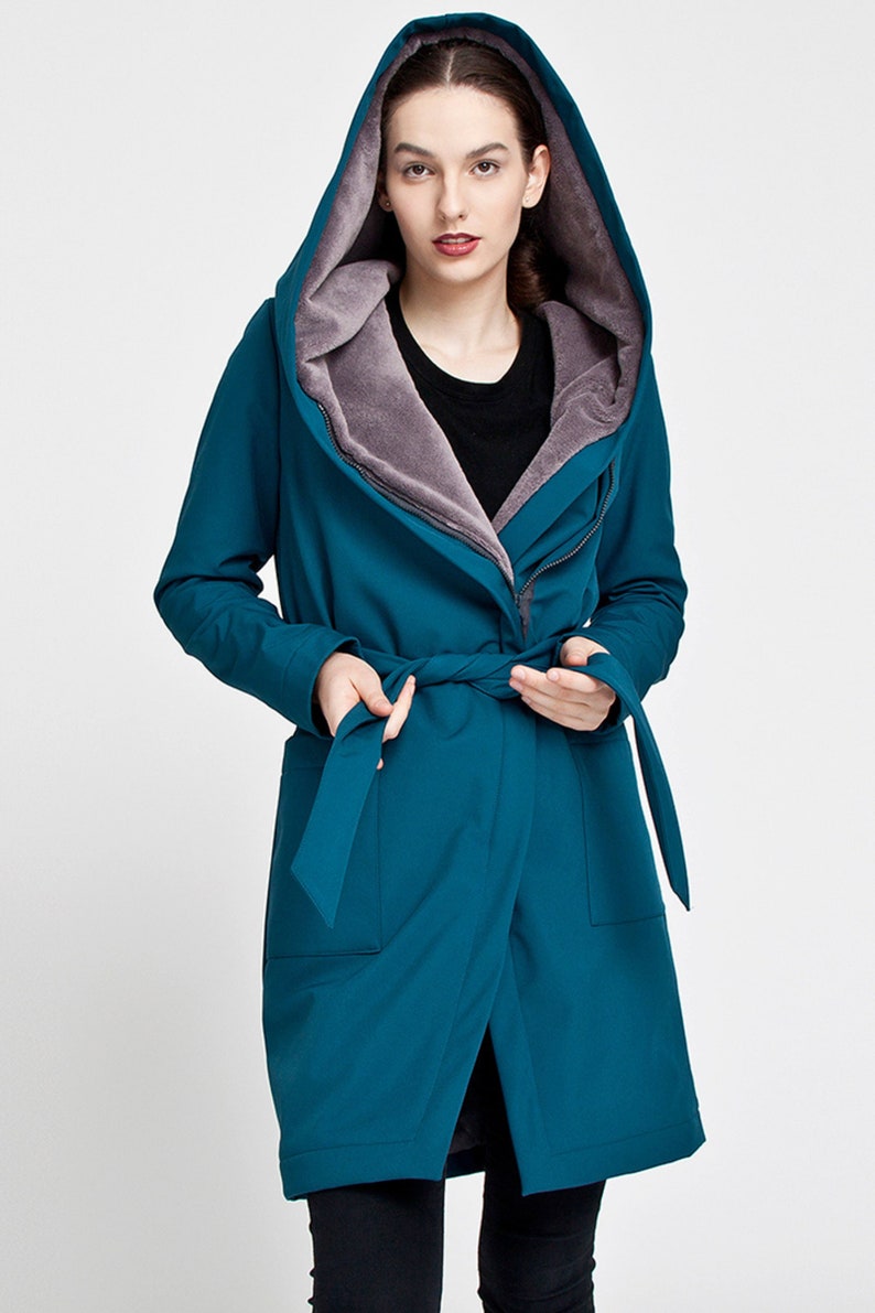 May include: A teal hooded coat featuring a soft grey faux fur lining, a wrap-style front with a matching fabric tie belt, long sleeves, and two front pockets. The coat has an oversized hood and a zipper closure partially visible under the wrap.