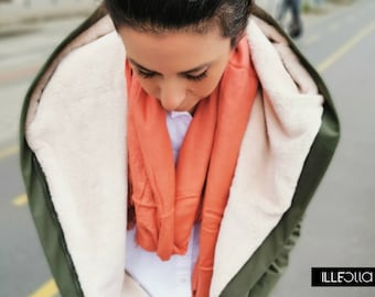 May include: A woman wearing a green hooded jacket with a white fleece lining and a coral scarf. The jacket has a zipper closure and a hood.
