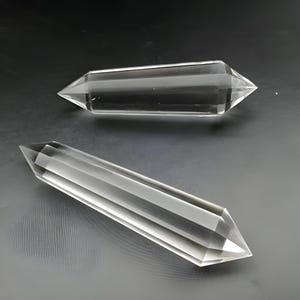 May include: Two clear quartz crystal wands with pointed ends. The wands are transparent and faceted, reflecting light. They are lying on a dark gray surface. The crystals are a spiritual or decorative item.