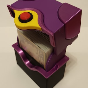 Könnte beinhalten: Ein lila-schwarzer Kartenhalter mit einem Stapel Karten darin. Der Halter hat ein einzigartiges Design mit gelbem Akzent und rotem Knopf. Der Halter ist aus Kunststoff und hat ein modernes Aussehen.