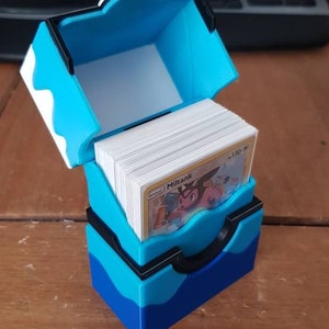 May include: A blue and black card storage box, open to reveal a stack of cards. The box is made of plastic and has a hinged lid. The cards are visible, with the word "Miltank" printed on one card.