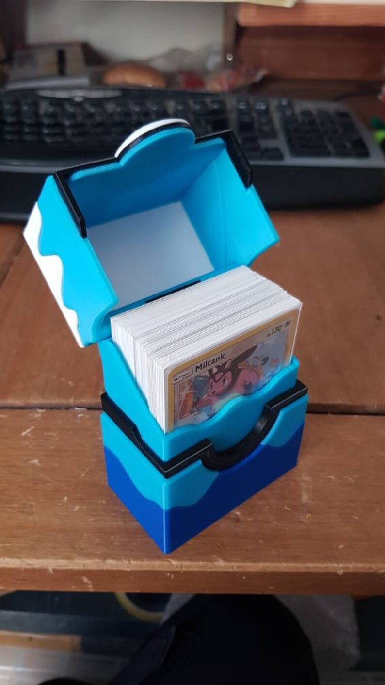 May include: A blue and black card storage box, open to reveal a stack of cards. The box is made of plastic and has a hinged lid. The cards are visible, with the word "Miltank" printed on one card.