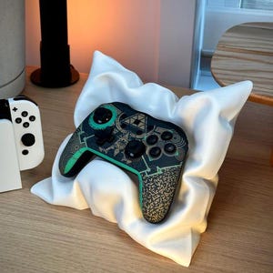 May include: A white, sculpted, pillow-shaped controller stand with a matte finish, holding a black wireless game controller. The controller features a detailed, intricate graphic design in shades of teal and gold, including geometric patterns and symbols.