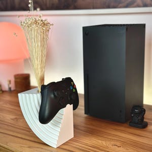 May include: A black Xbox Series X console and controller are displayed on a wooden surface. The controller rests on a white and pink decorative stand. A small black figurine and dried flowers are also present.