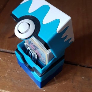 May include: A blue, white, and black card storage box, designed to resemble a stylized ball. The box is open, revealing a stack of cards inside. The box is made of plastic and sits on a wooden surface.
