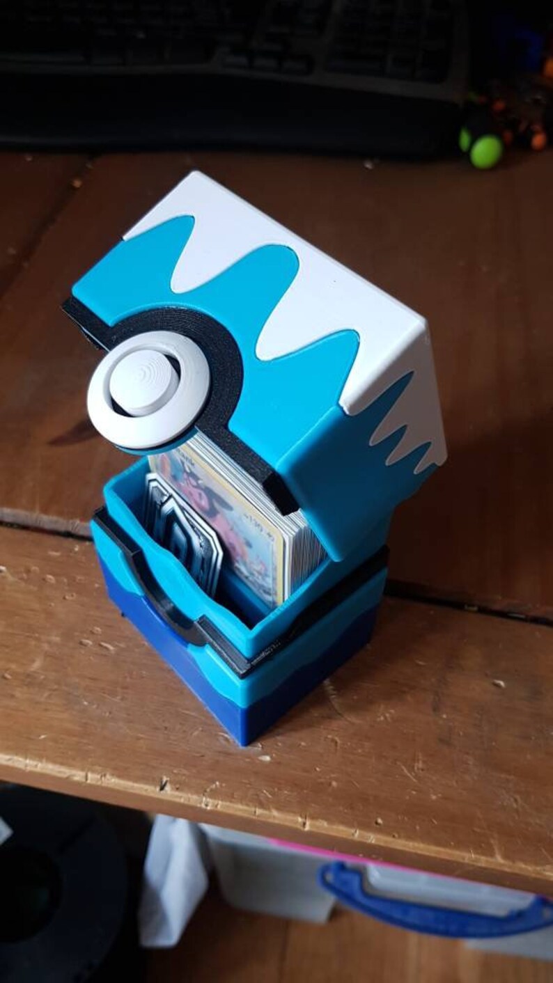 May include: A blue, white, and black card storage box, designed to resemble a stylized ball. The box is open, revealing a stack of cards inside. The box is made of plastic and sits on a wooden surface.