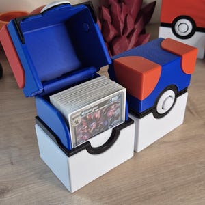 May include: A blue, red, and white card storage box, open to reveal a stack of cards. The box is designed to resemble a Pokeball. A second, closed Pokeball-themed box is visible, along with a red crystal sculpture.