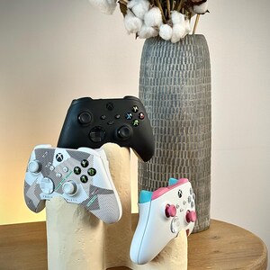 Può includere: Tre controller Xbox in nero, bianco e bianco con accenti rosa e blu sono esposti su un supporto color crema. Un vaso grigio alto e strutturato con steli di cotone è sullo sfondo, il tutto su un tavolo di legno.