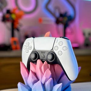 May include: A white and black wireless game controller resting in a 3D-printed stand shaped like a cluster of crystals. The crystal stand features a gradient color scheme transitioning from light pink at the top to light blue at the base.