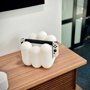 May include: A white, sculptural charging stand for a gaming console, featuring rounded, oblong shapes. The stand holds a black console with white controllers. The stand is on a wooden surface.