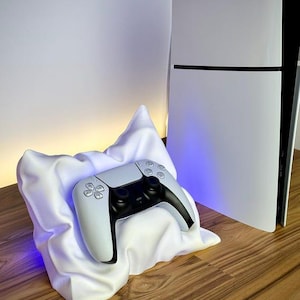 May include: A white gaming controller rests on a white, draped, pillow-like stand. Next to it stands a white and black gaming console featuring the PlayStation logo. The setup is illuminated by a soft blue light emanating from behind the console.