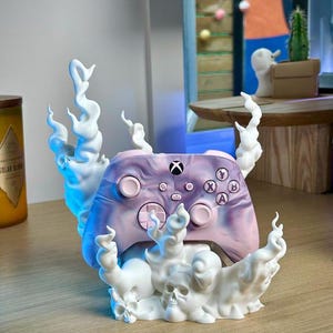 Può includere: Un controller wireless Xbox marmorizzato viola e rosa appoggiato su un supporto espositivo bianco stampato in 3D. Il supporto presenta volute di fumo bianco scolpite e piccoli dettagli a forma di teschio alla base per sostenere il controller.