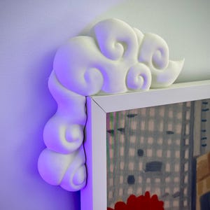 Op de afbeelding: Een wit, wolkvormig decoratief element bevestigd aan de hoek van een ingelijst kunstwerk. De wolk heeft een gestructureerd, driedimensionaal ontwerp met wervelende details. Het kunstwerk toont een stadsgezicht met rode en grijze tinten.