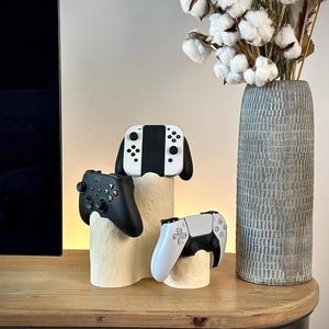 Op de afbeelding: Een crèmekleurige standaard met meerdere niveaus en een getextureerd oppervlak houdt drie videogamecontrollers vast: een wit-zwarte handheld consolecontroller bovenaan, een zwarte controller links en een wit-zwarte controller rechts.